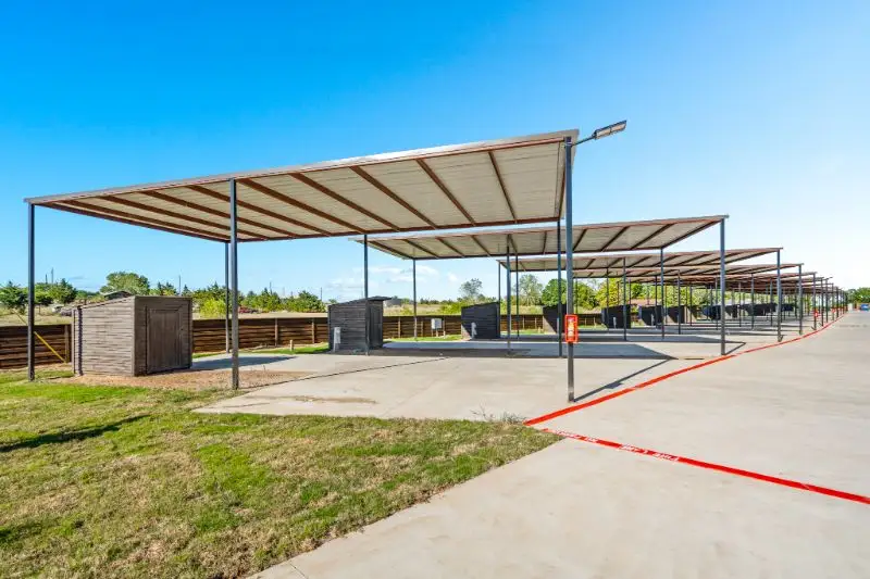 Row of metal covered RV parking canopies over concrete pads, with storage sheds and wooden fencing along the grassy